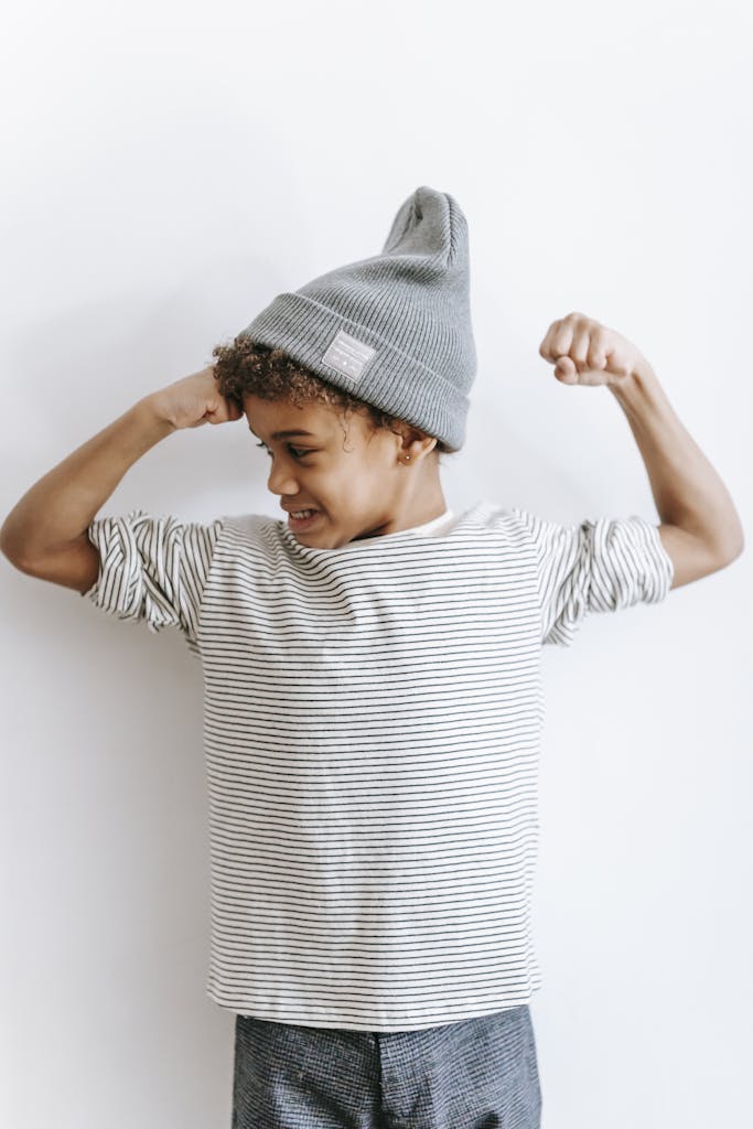 Positive African American boy in trendy hat and striped outfit showing strength while standing against white background in light room