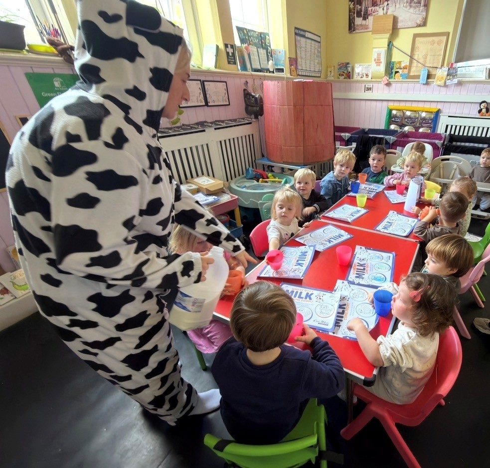 A member of staff in a cow onesie pouring milk for nursery children at their tables